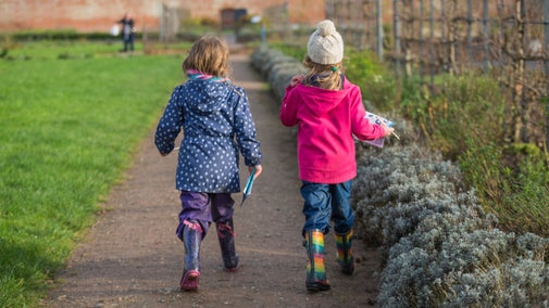 Two children wearing winter coats and hat walk through a walled garden, carrying a trail activity sheet and pencils